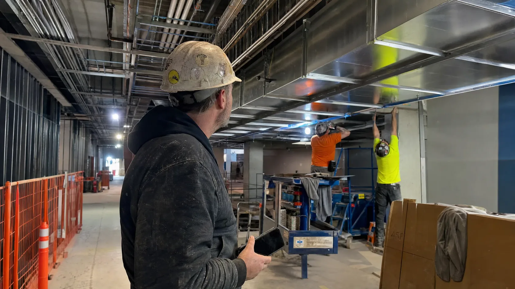A tradesman on an active jobsite checking his phone beneath ductwork being installed overhead