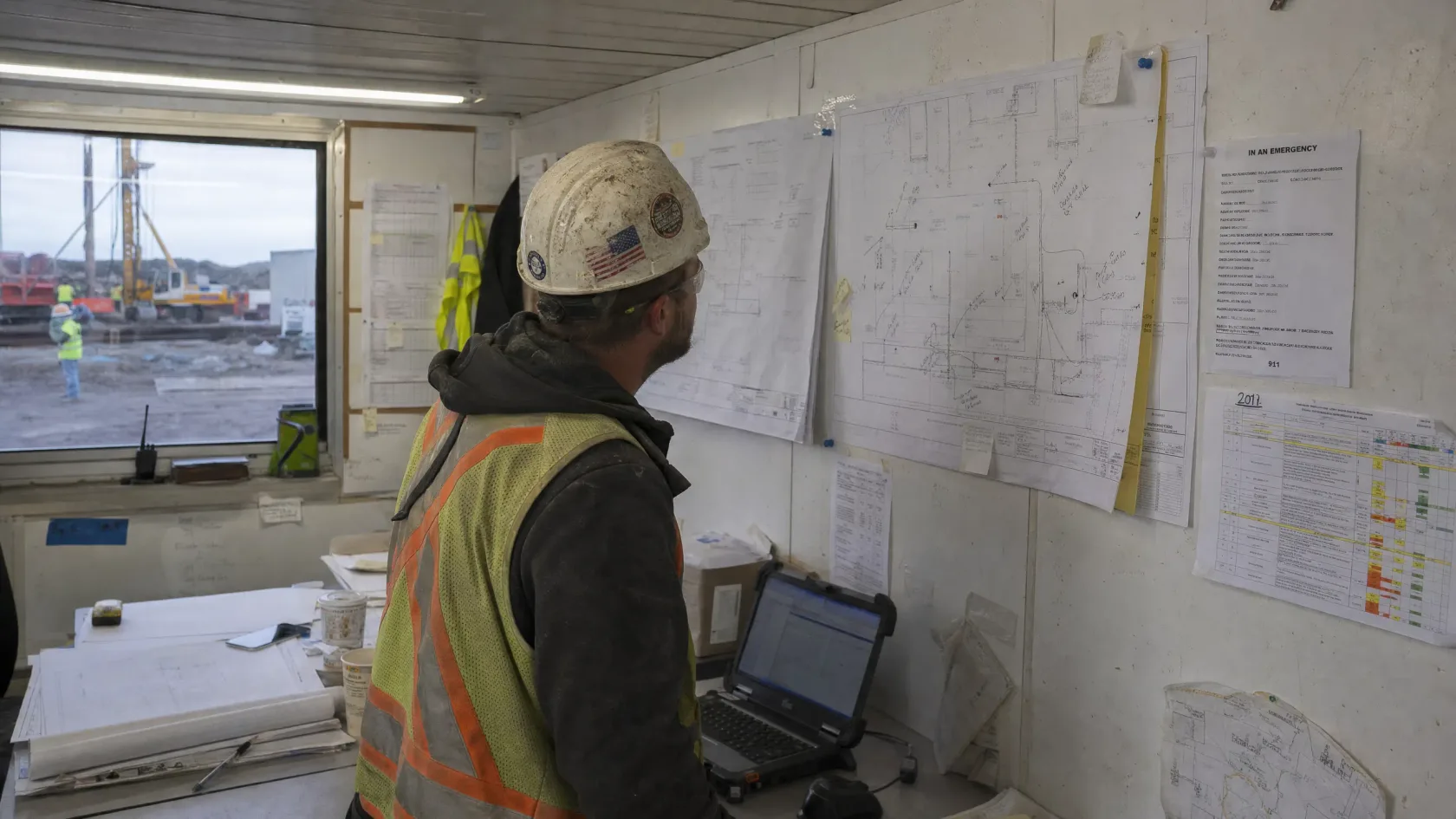 A superintendent working at a laptop in a site trailer, marked-up drawings and schedules taped to the wall beside him